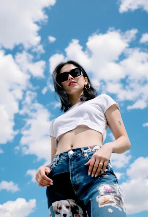 A young woman with dark hair and rectangular black sunglasses, wearing a white crop top and patchwork, photo-printed blue jeans, stands against a bright blue sky with scattered white clouds. The camera angle is low, looking up towards her. She holds the waistband of her jeans. A small flower tattoo is visible on her left upper arm. She wears a simple gold ring on her left ring finger.  The overall style is casual and trendy with a slightly retro feel. The image is well-lit, likely with natural daylight, and has a cool, almost nostalgic tone.